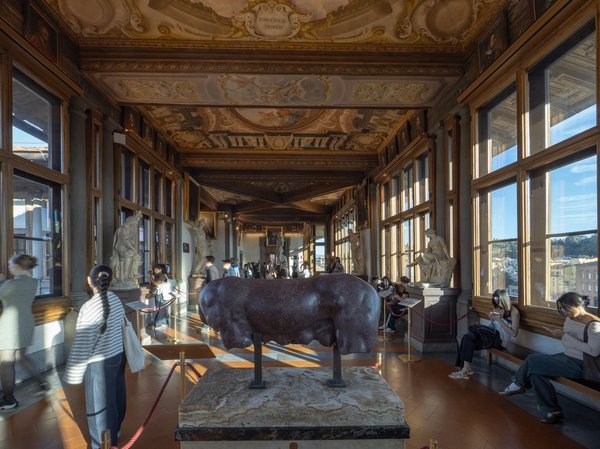 Corridor of the Uffizi Gallery in Florence with statues, a frescoed ceiling, and windows overlooking the city.