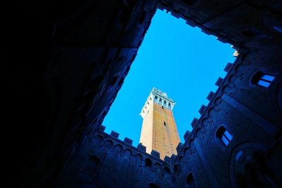 Low-angle view of the Torre del Mangia in Siena, framed by historic buildings and a deep blue sky.