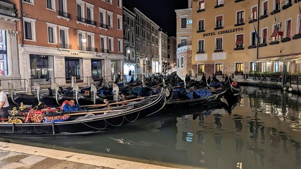 Una fila di gondole ormeggiate lungo un canale di Venezia di notte, illuminate dai lampioni.