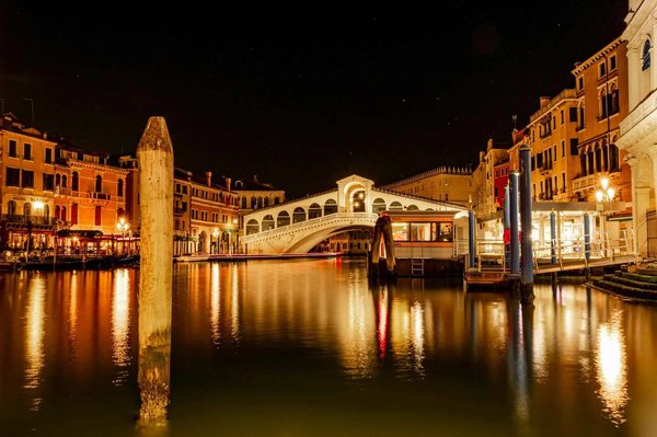 Ponte di Rialto illuminato di notte a Venezia con riflessi sull'acqua.