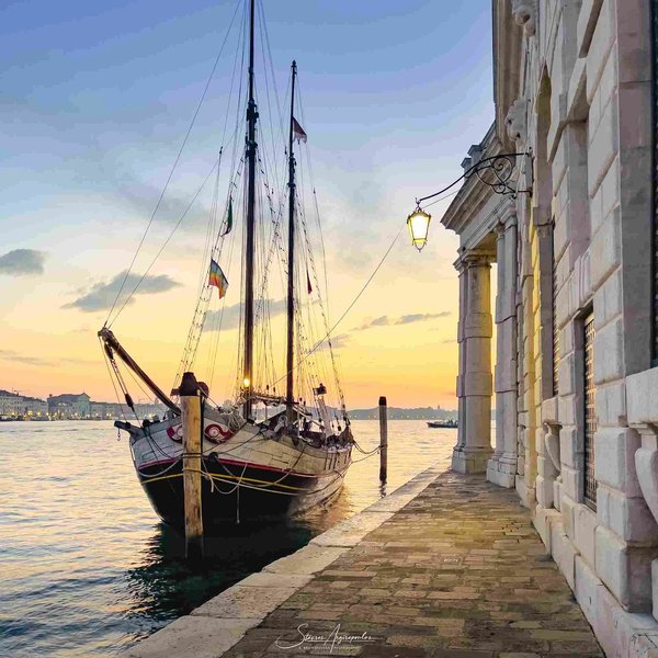 Sailboat moored along the Riva degli Schiavoni in Venice at sunset, with historic buildings and a lit lamppost.
