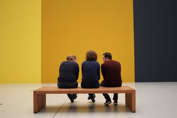 Tre persone sedute su una panchina di legno davanti a un muro giallo e nero al MO Museum di Vilnius.
