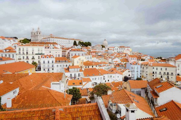 Panoramic view of red rooftops and historic streets of the Alfama neighborhood in Lisbon, with a cloudy sky in the background.