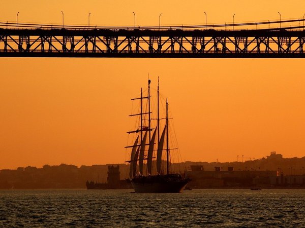 Sunset over the Tagus River in Lisbon with the 25 de Abril Bridge and a sailboat in the foreground.