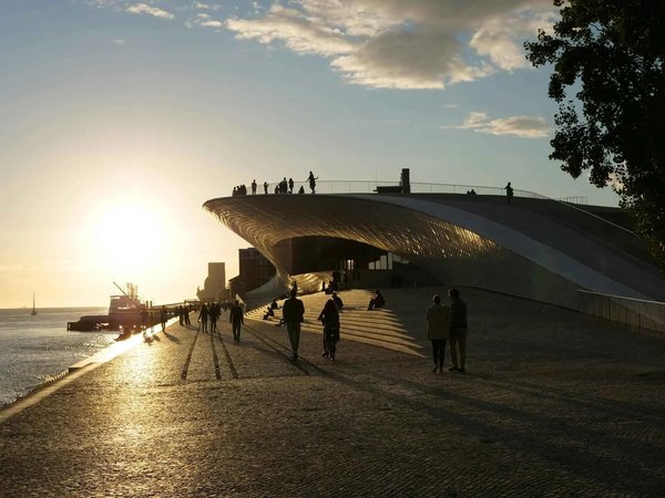 Sunset at MAAT in Lisbon with people walking along the river and the iconic modern building illuminated by the sun.