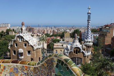 View of Park Güell in Barcelona with iconic buildings and colorful mosaics, with the city skyline and the sea in the background.