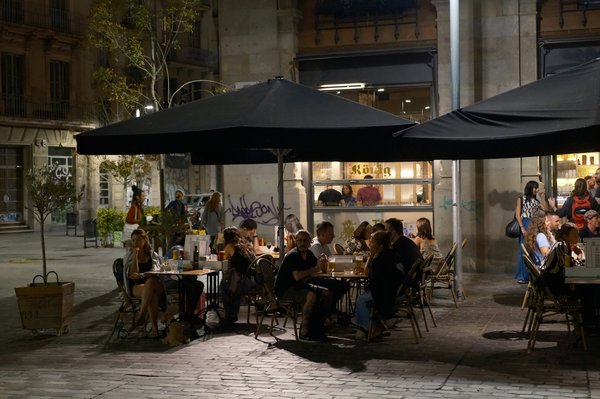 People dining at outdoor terrace tables under umbrellas on a lively plaza at night.