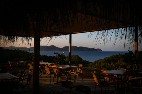 An empty seafront restaurant terrace under a thatched roof, set with tables overlooking the coastline at dusk.