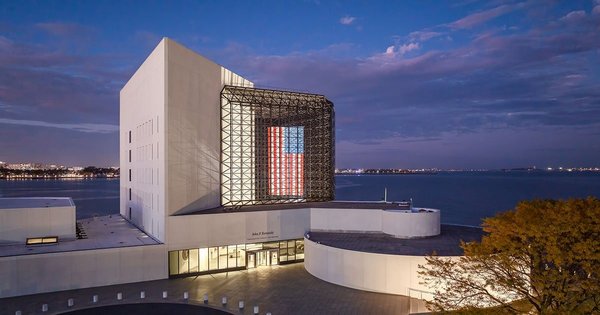 John F. Kennedy Presidential Library in Boston at sunset, overlooking the ocean with an illuminated American flag.