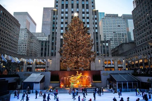 Albero di Natale illuminato al Rockefeller Center con pista di pattinaggio e skyline di New York sullo sfondo.