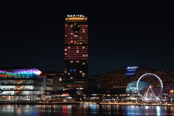 Darling Harbour di notte, con la ruota panoramica illuminata e le torri Sofitel e Novotel riflesse nell'acqua.