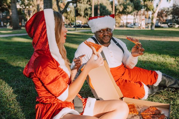 Una coppia in costume da Babbo Natale condivide un picnic a base di pizza sull'erba in un parco soleggiato, Natale in stile australiano.