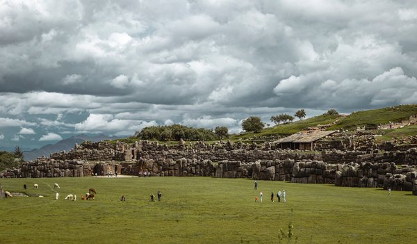 Rovine di Sacsayhuaman a Cusco, Perù, con prati verdi e cielo nuvoloso, visitatori e lama sullo sfondo.